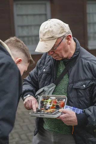 Ein Eberhard Scheibe, ein älterer Mann, zeigt einem jüngeren eine Box mit bunten Fliegenködern fürs Fliegenfischen. Beide schauen aufmerksam auf die Auswahl.