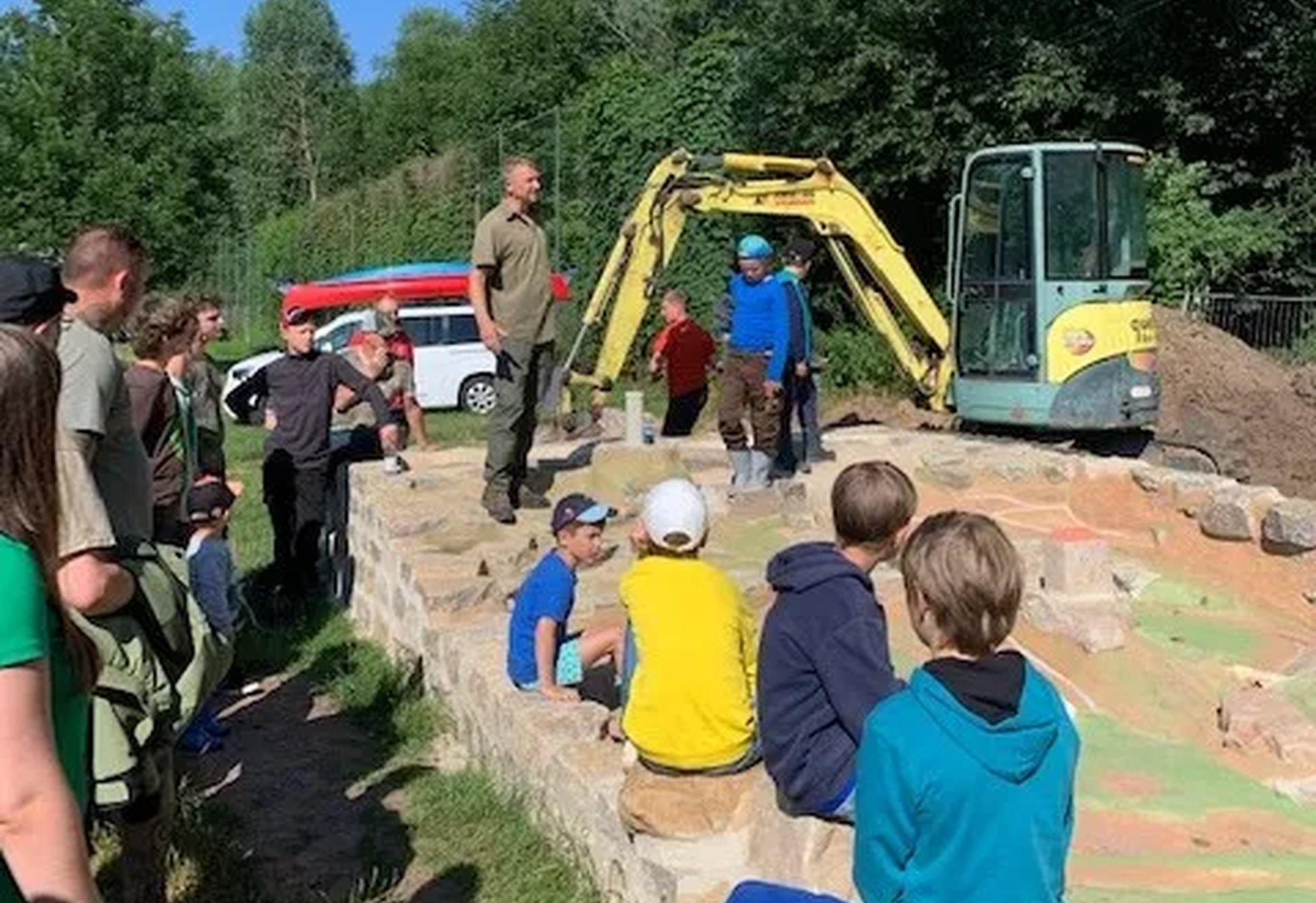 Eine Gruppe von Kindern sitzt und steht um ein sandiges Areal mit einem kleinen Bagger, während ein Erwachsener spricht. Im Hintergrund sind Bäume und geparkte Autos zu sehen.
