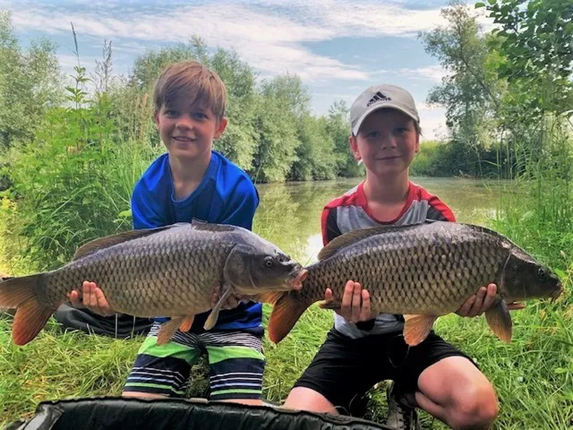 Zwei Jungen sitzen am Ufer eines Sees und halten jeweils einen großen Fisch in den Händen.
