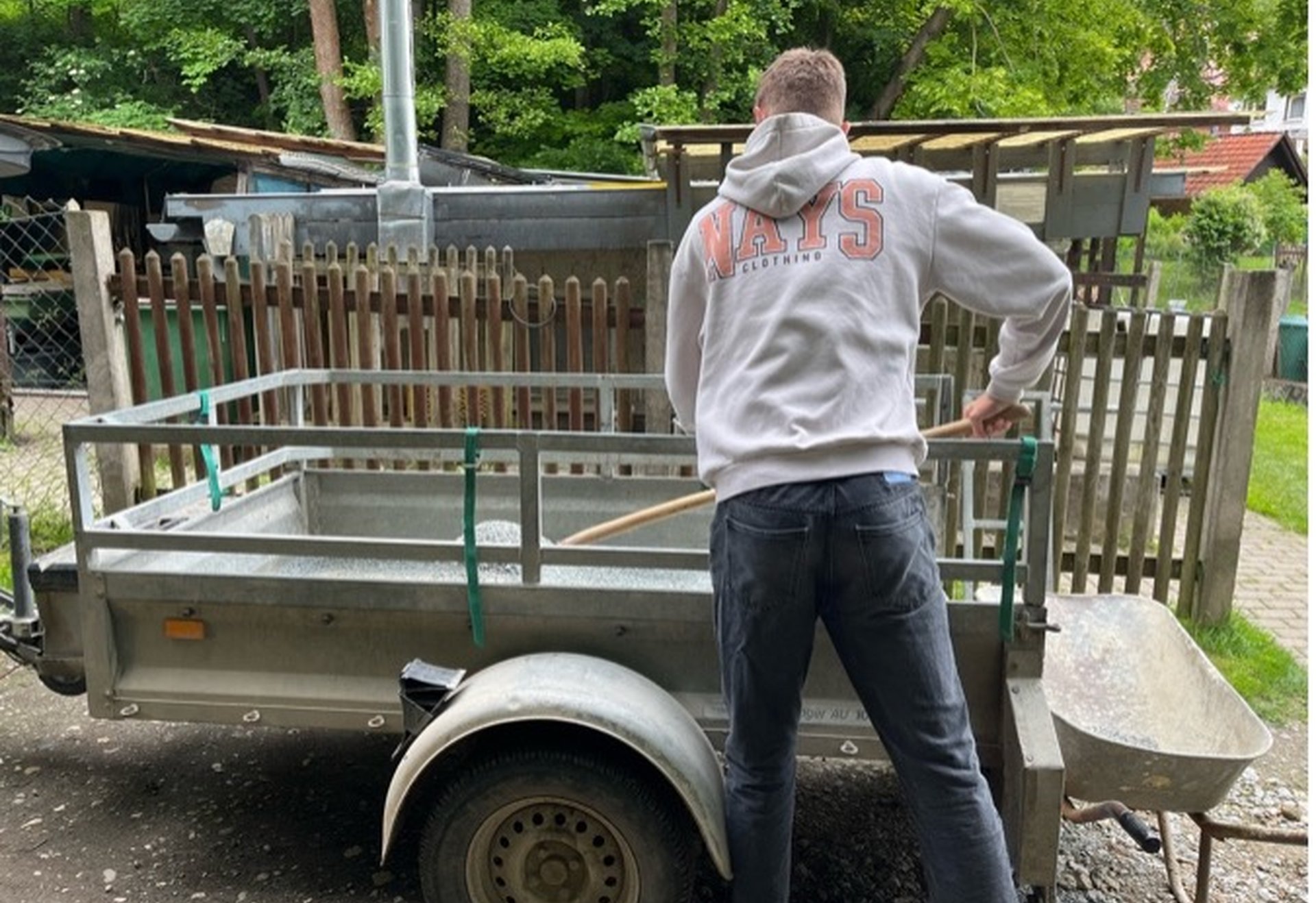 Ein Mann steht an einem Anhänger und entlädt mit einer Schaufel Material in eine Schubkarre.