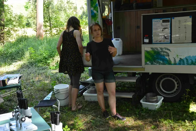 Zwei Frauen stehen neben einem mobilen Laborfahrzeug im Grünen, umgeben von naturwissenschaftlicher Ausrüstung wie Mikroskopen und Eimern.