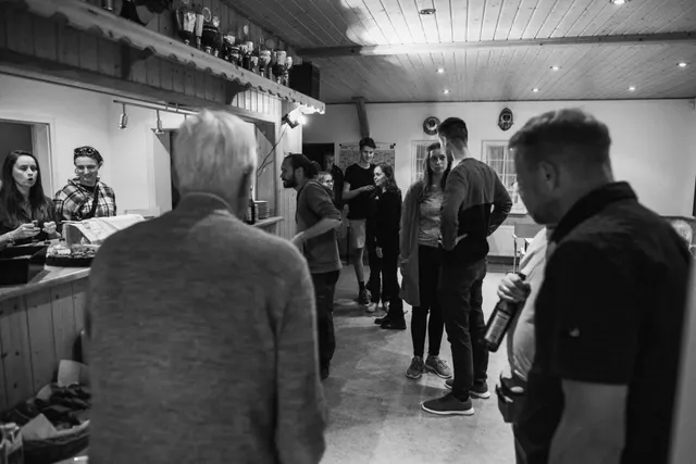 Mehrere Menschen stehen und unterhalten sich in einem Raum, einige warten an einer Theke; die Szene ist in Schwarz-Weiß gehalten.