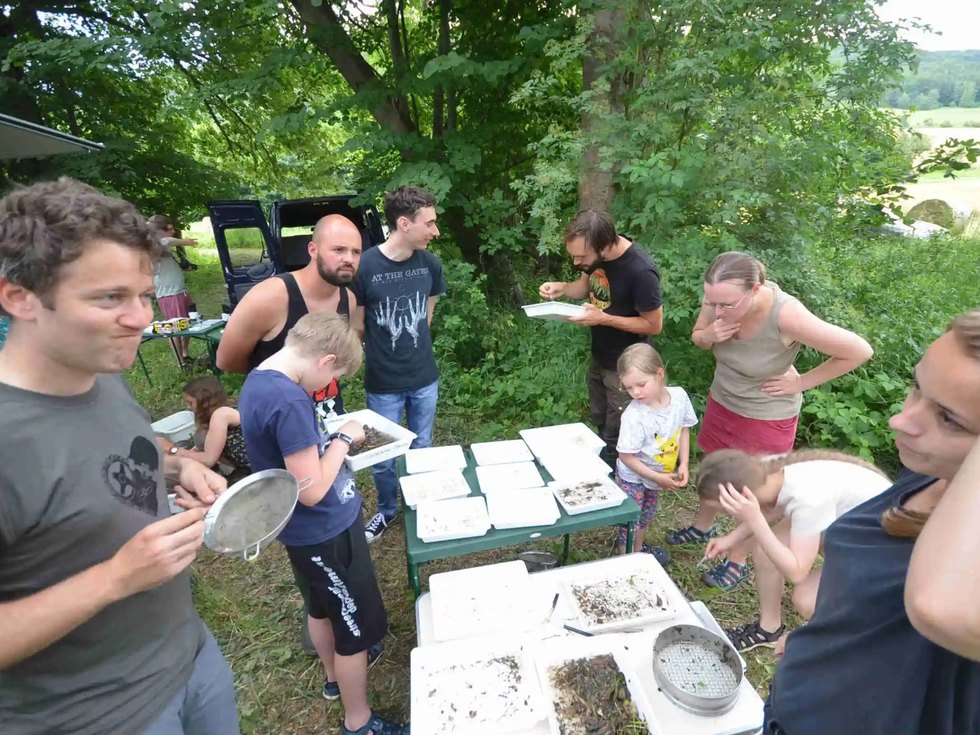 Mehrere Erwachsene und Kinder stehen im Freien um Tische mit verschiedenen Schalen und Behältern, untersuchen den Inhalt und unterhalten sich. Im Hintergrund sind Bäume und Wiese zu sehen.