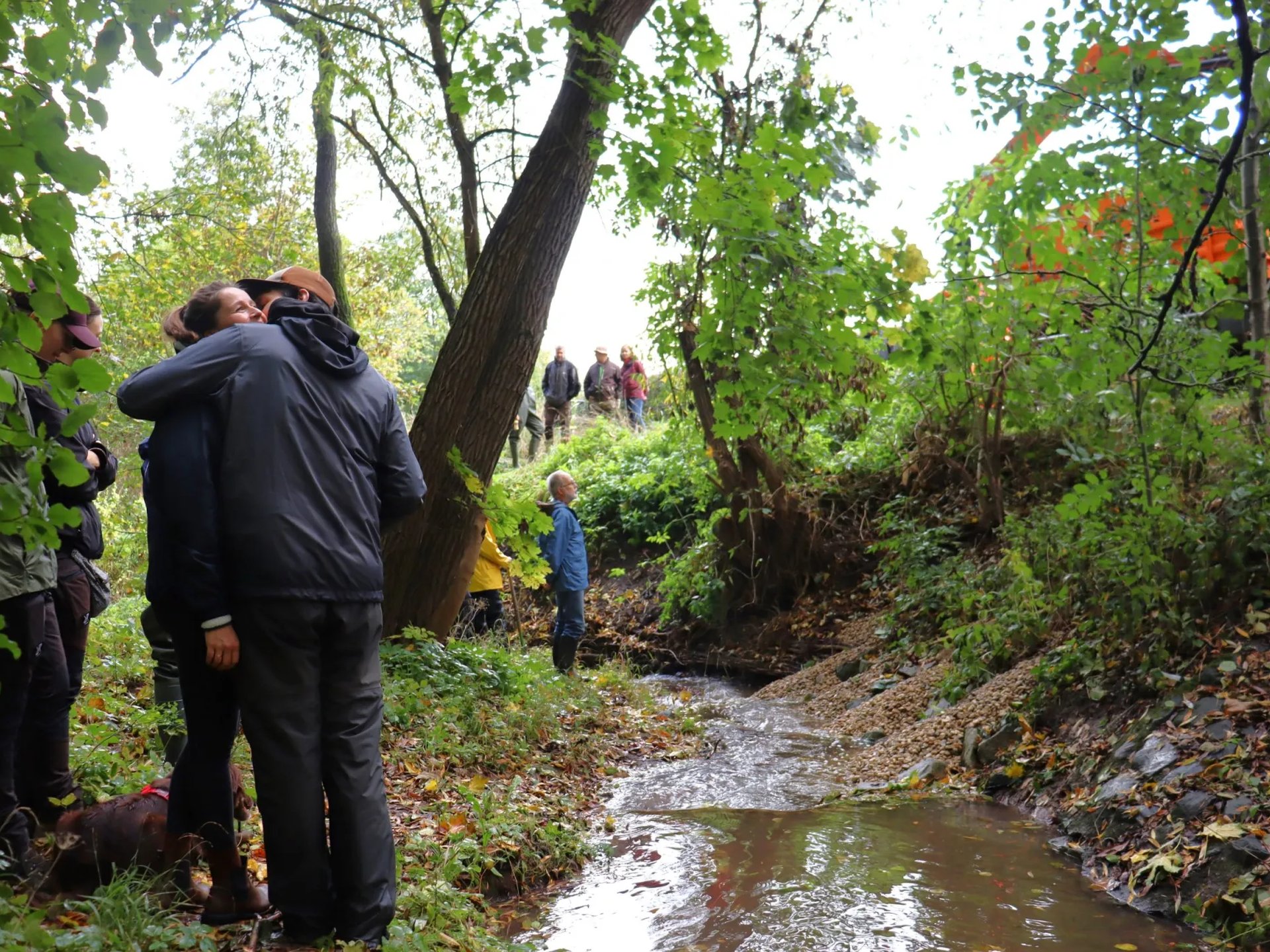 Mehrere Menschen stehen an einem kleinen Bach im Wald, einige umarmen sich, andere beobachten die Umgebung.