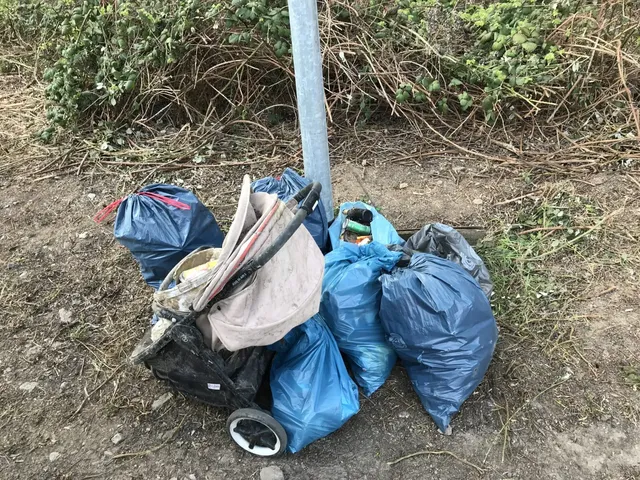Mehrere blaue Müllsäcke, ein Einkaufswagen mit Müll und verschiedene Abfälle liegen am Wegesrand vor einer Böschung.