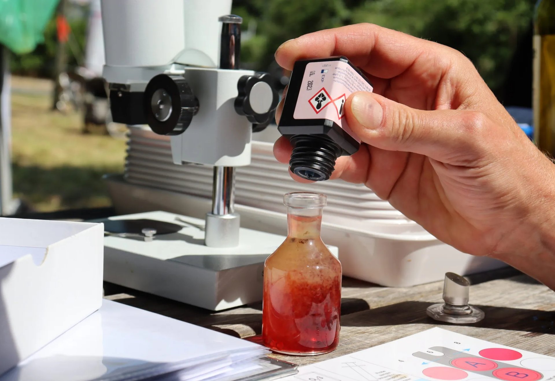 Eine Hand gießt eine Chemikalie aus einer Flasche in ein Glasgefäß vor einem Mikroskop.