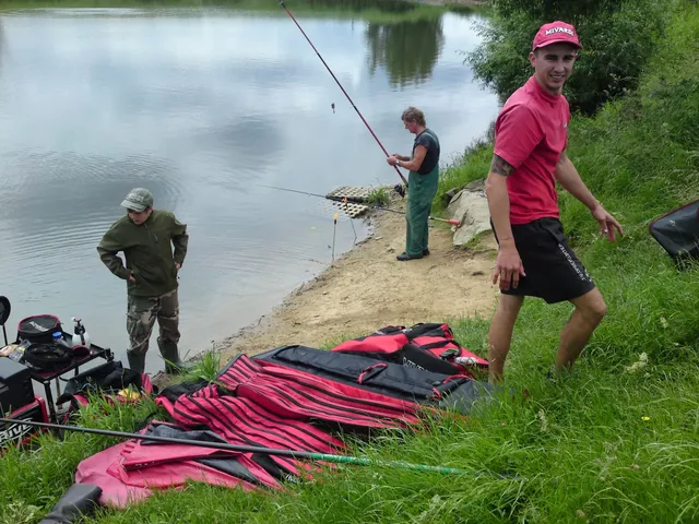 Drei Männer angeln an einem Seeufer, einer steht im Vordergrund auf einer Wiese neben zusammengelegtem Angelausrüstungs-Taschen.