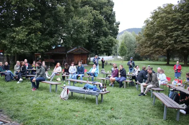 Mehrere Menschen sitzen auf Bänken und Tischen im Freien auf einer Wiese, umgeben von Bäumen.