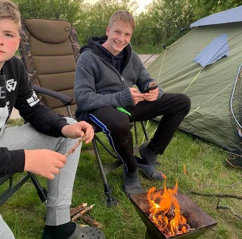 Zwei Jungen sitzen beim Camping auf Stühlen neben einem Zelt und grillen am offenen Feuer Marshmallows.