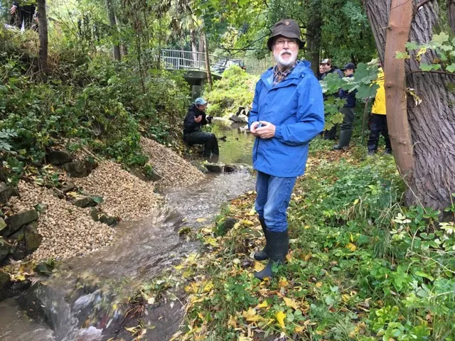 Ein Mann steht in Gummistiefeln und blauer Jacke an einem kleinen Bach, während mehrere weitere Personen am Ufer und im Wasser arbeiten, umgeben von dichtem Grün.