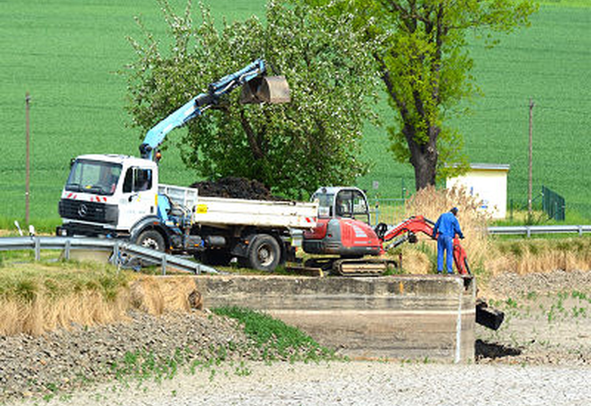 Ein Bagger und ein Lkw arbeiten an einer Baustelle neben einem trockenen Flussbett, ein Arbeiter steht daneben.