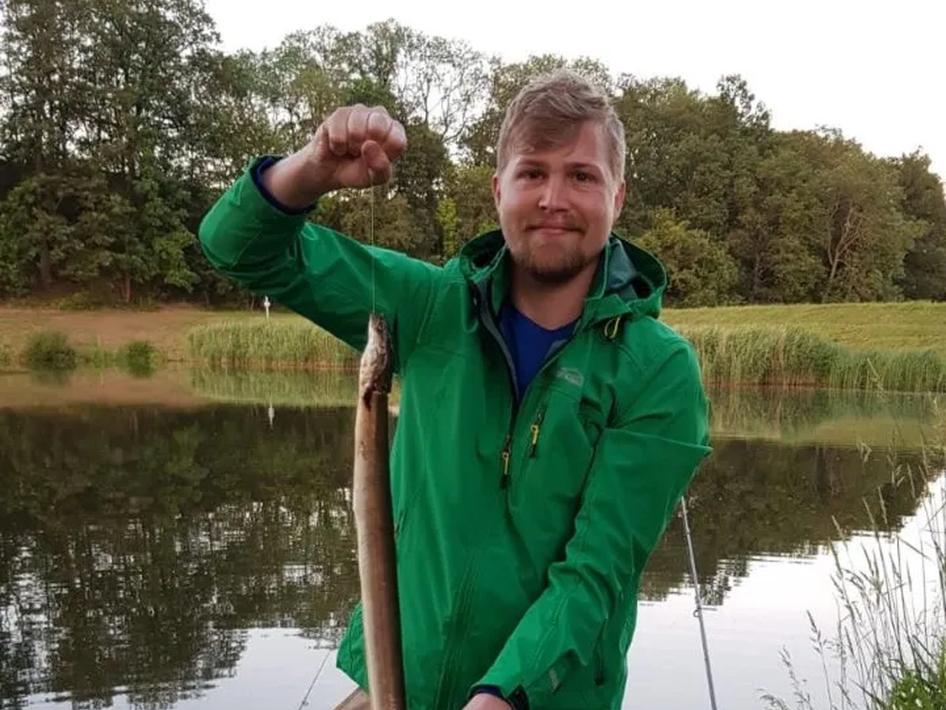 Ein Mann steht am Flussufer und hält einen langen Fisch in der Hand, im Hintergrund Angelruten und Wasser.