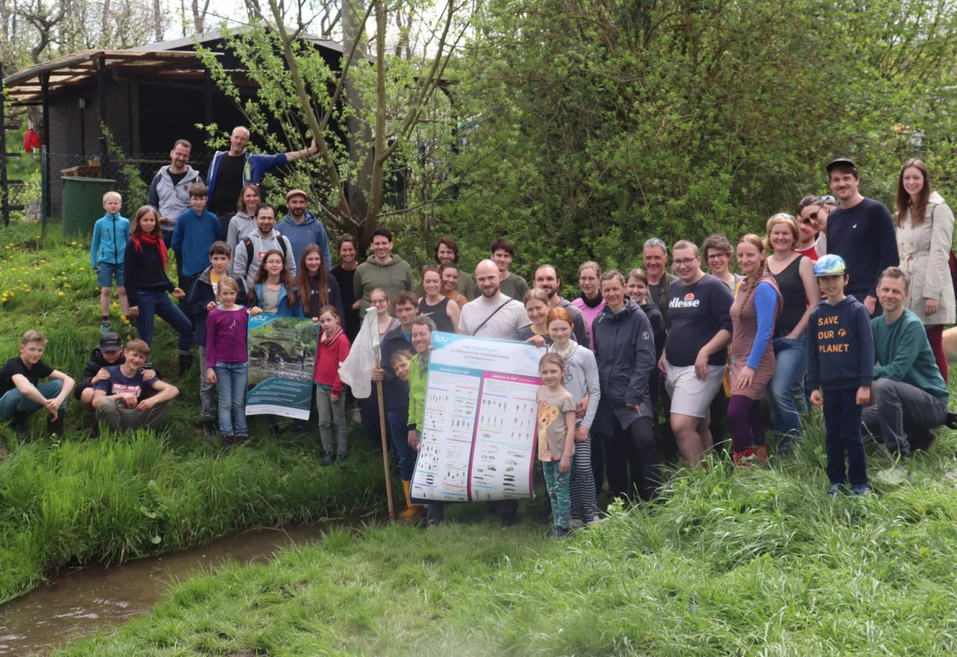 Eine große Gruppe von Menschen steht und sitzt im Grünen an einem Bach, sie posieren gemeinsam für ein Gruppenfoto, einige halten ein großes Plakat.