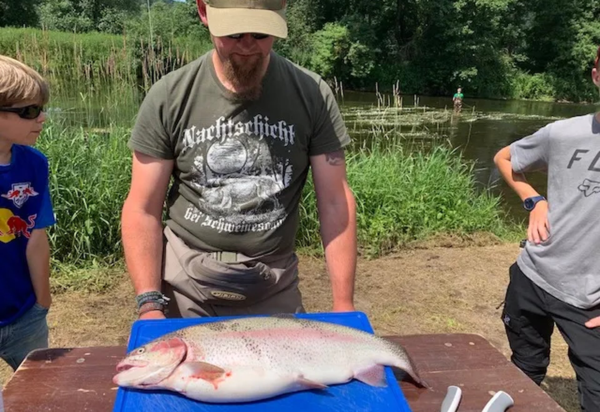 Ein Mann steht an einem Tisch im Freien und präsentiert eine große Forelle auf einem blauen Brett, daneben stehen zwei Kinder und im Hintergrund ist ein Teich zu sehen.