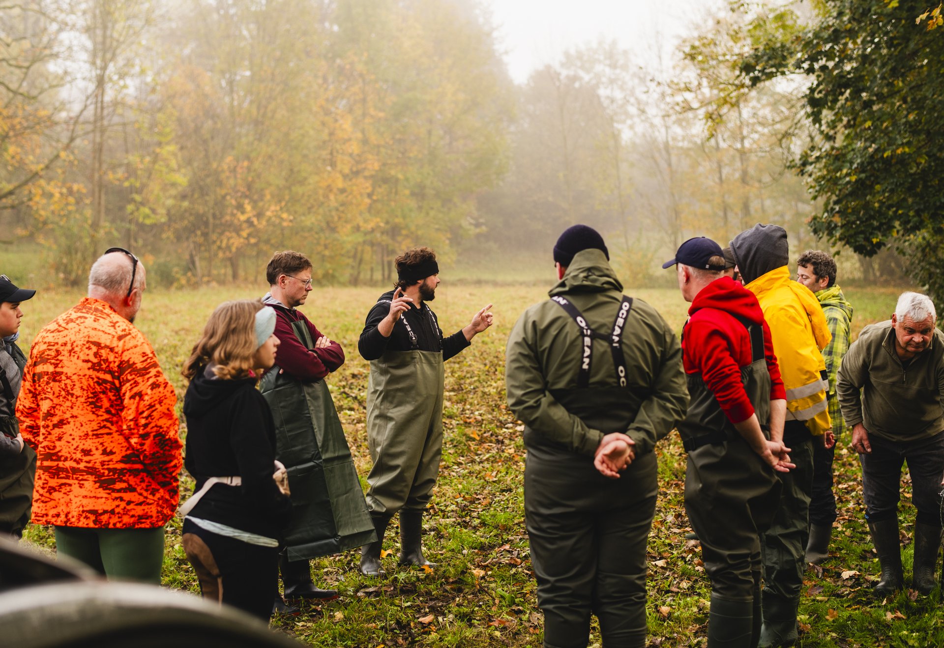 Eine Gruppe von Menschen steht im Freien auf einer Wiese im Herbst und hört einer Person in der Mitte aufmerksam zu.