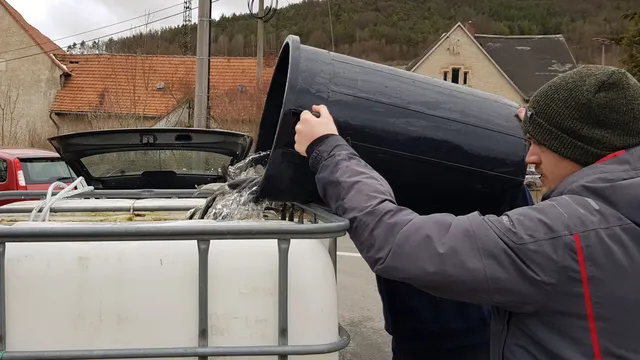 Ein Mann gießt Wasser aus einem großen schwarzen Behälter in einen weißen Wassertank draußen.