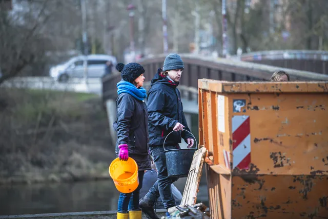 Zwei Personen werfen Müll in einen großen Container im Freien, beide tragen Winterkleidung und Gummistiefel.