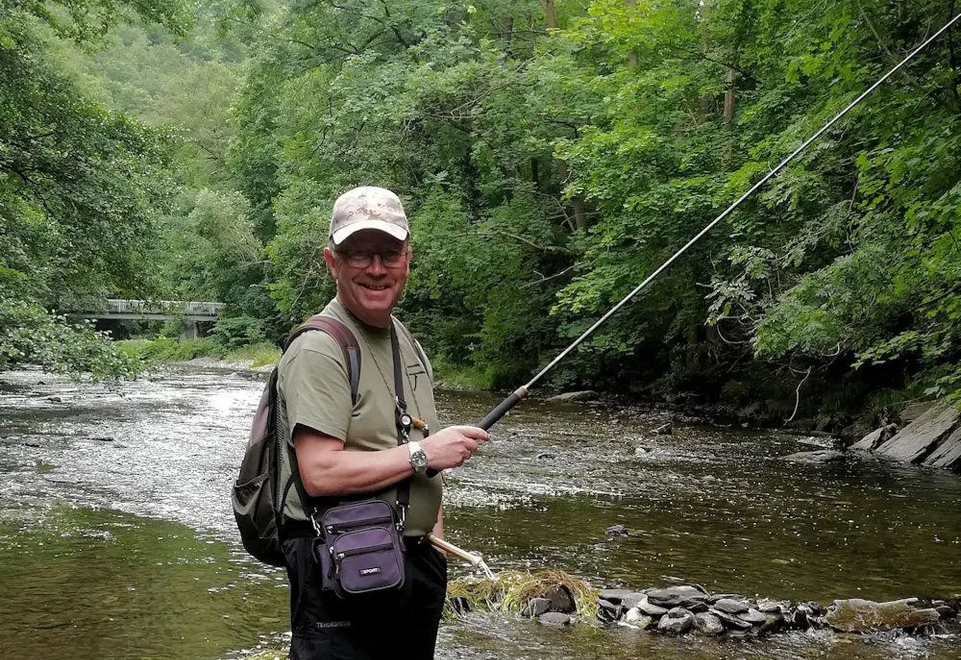 Ein Mann steht mit Angelrute an einem Fluss, umgeben von grüner Natur.