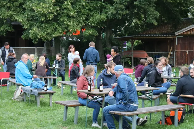 Mehrere Menschen sitzen und stehen an Biertischgarnituren im Freien und unterhalten sich.