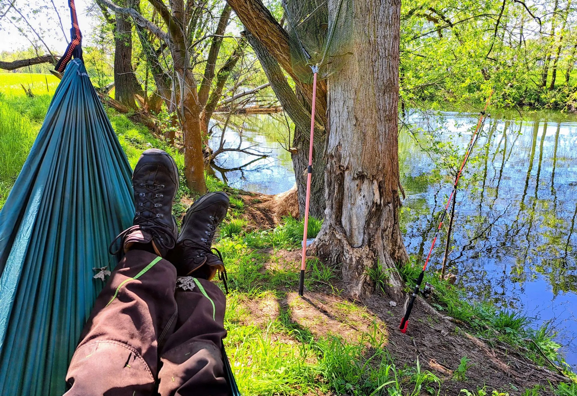 Person liegt in einer Hängematte am Flussufer, mit Blick auf zwei aufgestellte Angelruten. Sonnenlicht fällt durch die Bäume auf das ruhige Wasser.