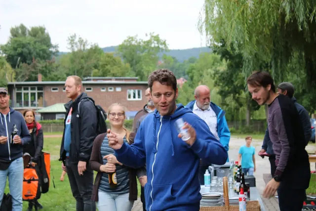 Mehrere Menschen stehen im Freien zusammen, einige von ihnen halten Getränke, im Vordergrund steht ein Mann in blauer Jacke und lächelt in die Kamera. Im Hintergrund sieht man Bäume und ein Gebäude.