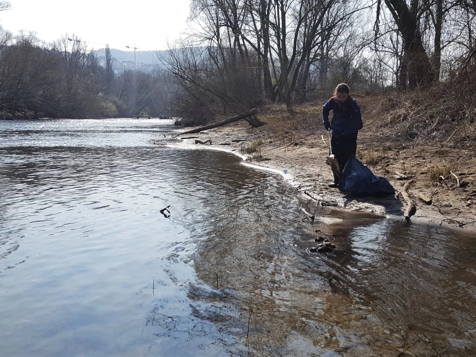 Eine Person steht am Ufer eines Flusses und sammelt Müll in einem blauen Sack; Bäume und kahles Gestrüpp säumen das Ufer.