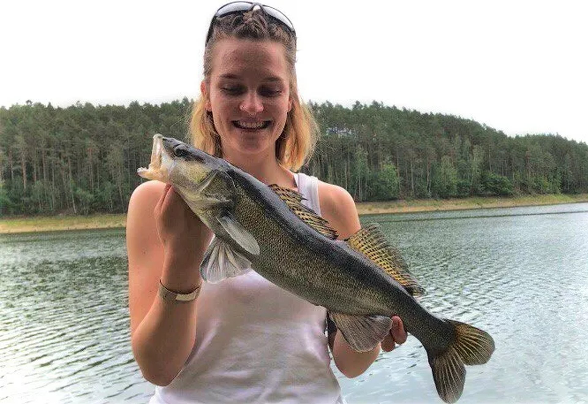 Eine junge Frau hält am Ufer eines Sees stolz einen großen Fisch in den Händen.