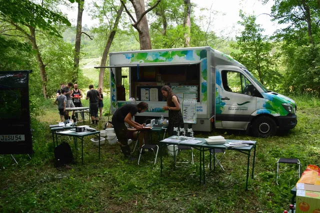 Ein mobiler Laborwagen steht in einer Waldlichtung, mehrere Menschen arbeiten an Tischen mit wissenschaftlichen Geräten im Freien.