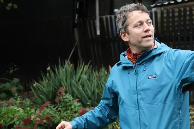 Ein Mann in blauer Jacke steht im Garten, im Hintergrund sind grüne Pflanzen und ein dunkler Zaun zu sehen.