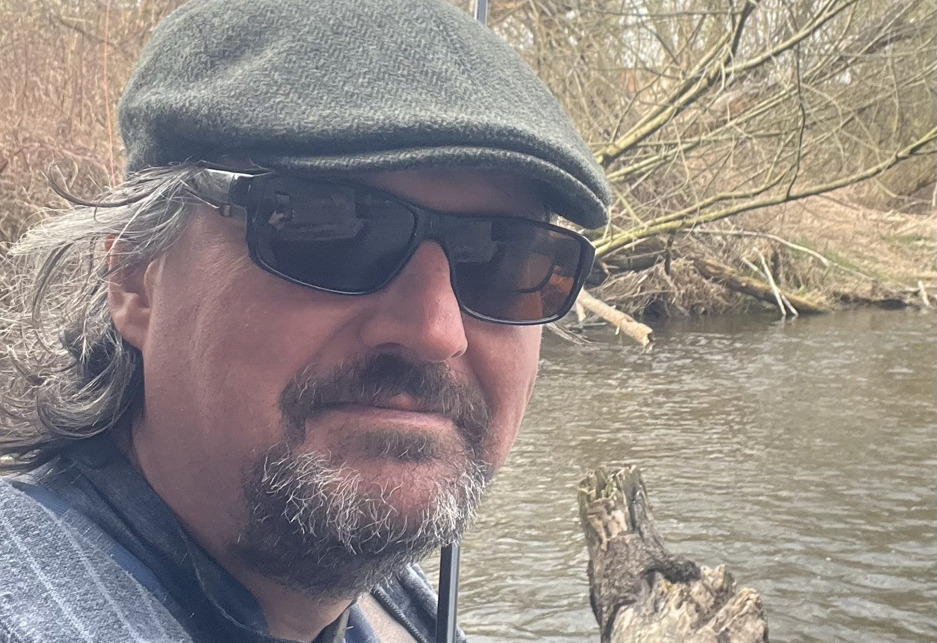 Ein Mann mit Sonnenbrille und Schirmmütze macht ein Selfie am Flussufer. Er trägt Angelkleidung und hält eine Fliegenrute, im Hintergrund sind Wasser, Äste und ein Baumstamm im Fluss zu sehen.