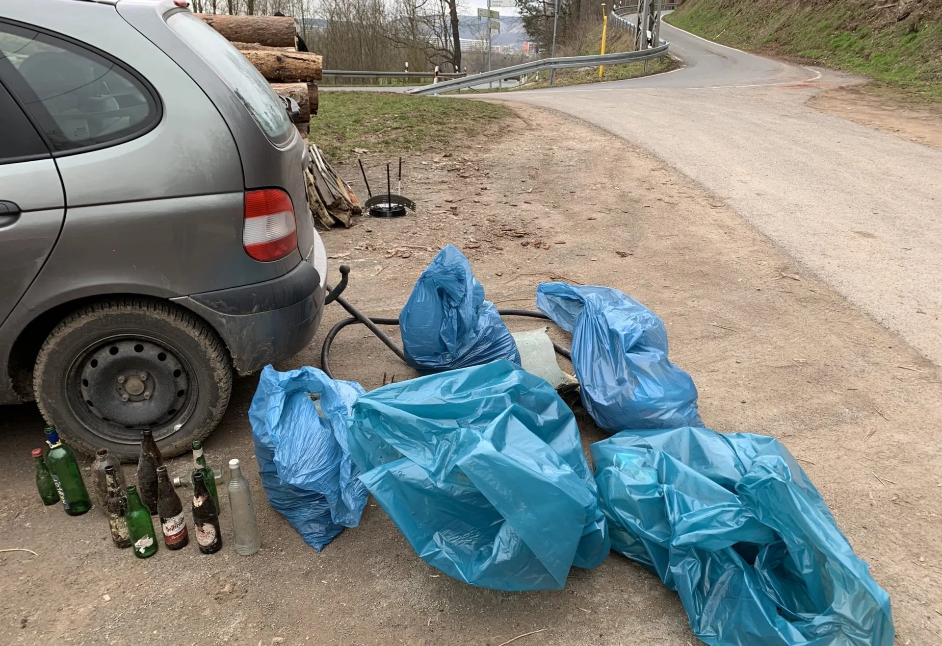 Mehrere blaue Müllsäcke und leere Flaschen stehen neben einem Auto am Straßenrand.