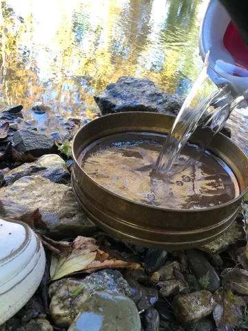 Wasser wird aus einem Krug in ein Metallsieb am Ufer eines Flusses gegossen.