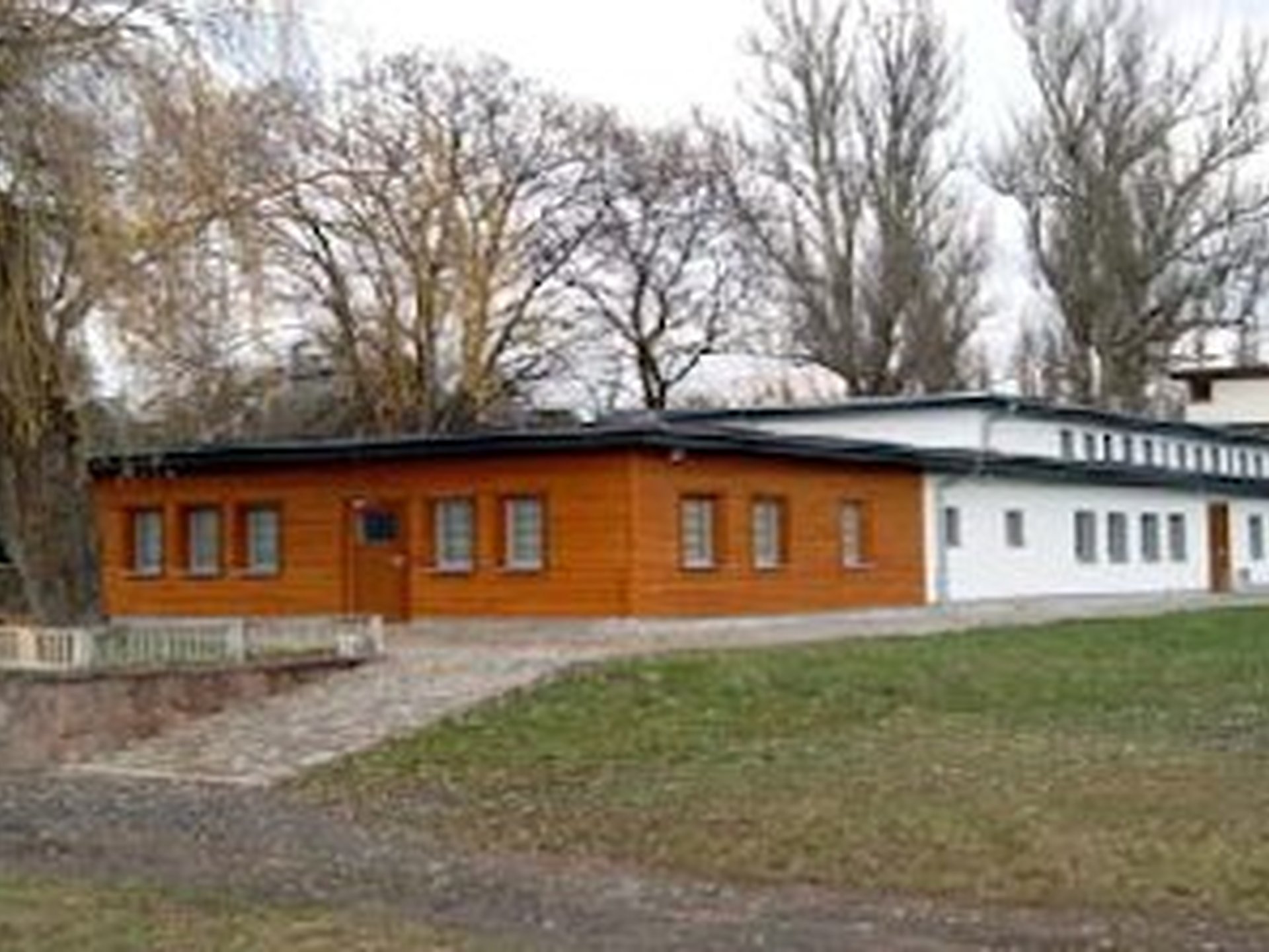 Flaches Gebäude mit Holz- und weißer Fassade, umgeben von Bäumen und Wiese.