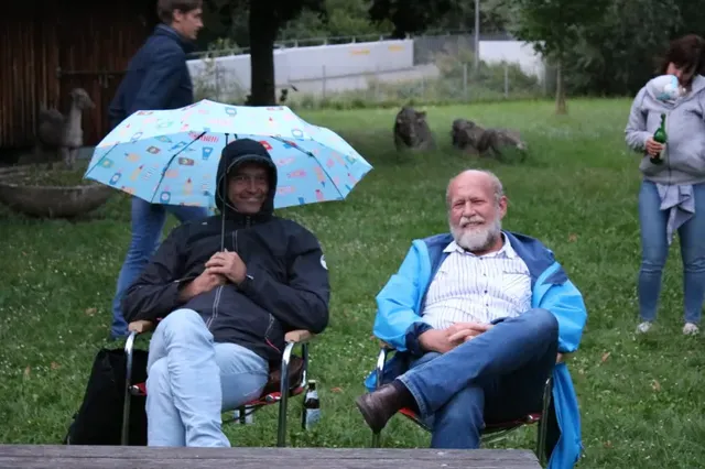Zwei Männer sitzen auf Stühlen im Freien; einer hält einen bunten Regenschirm, der andere lächelt. Im Hintergrund stehen weitere Personen auf einer Wiese.