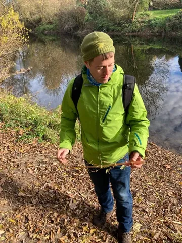 Eine Person mit grüner Jacke und Mütze steht am Ufer eines Flusses, hält einen Zweig und schaut nach unten.