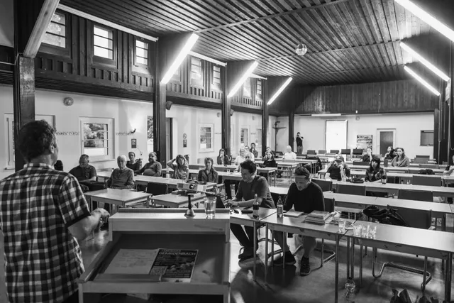 Schwarzweiß-Foto eines Seminarraums, in dem ein Vortragender vor mehreren sitzenden Zuhörern spricht.