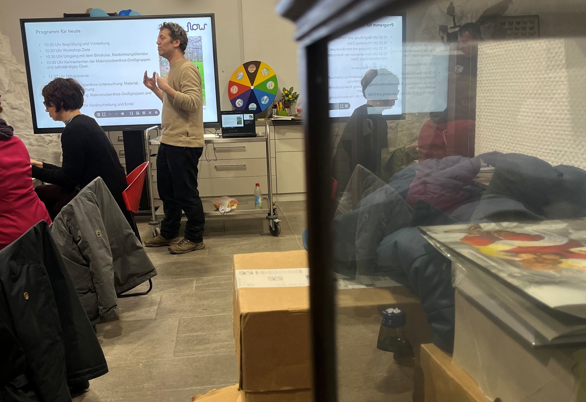 Ein Mann hält einen Vortrag vor einer kleinen Gruppe in einem Seminarraum, im Hintergrund steht ein Bildschirm mit einer Präsentation.