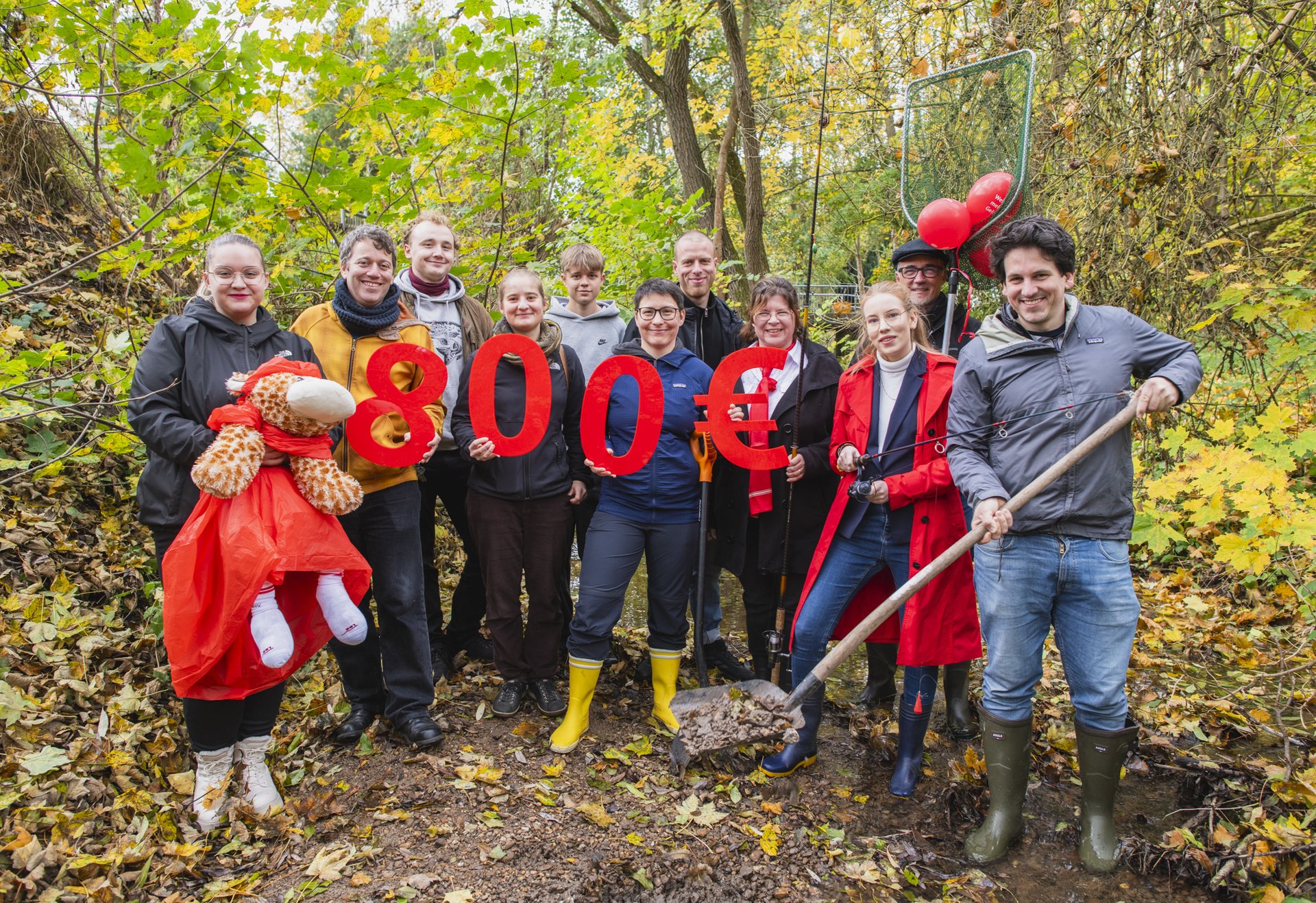 Eine Gruppe von Menschen steht im herbstlichen Wald und hält große rote Zahlen und ein Eurozeichen, die zusammen „800 €“ ergeben; vorne rechts hält ein Mann einen Spaten in den Händen.
