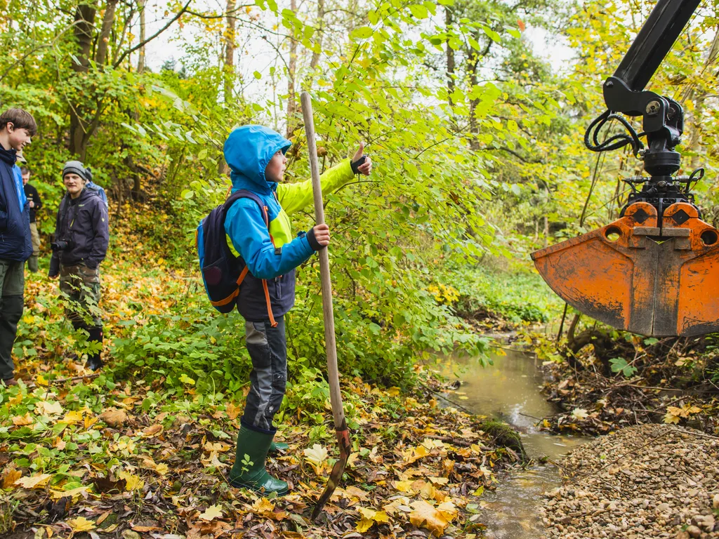 Eine Gruppe von Personen steht an einem kleinen Bach im Wald. Im Vordergrund hält ein Kind einen Holzstab und zeigt mit dem Daumen nach oben, während ein Baggerarm Kies in das Gewässer einbringt. Herbstliches Laub bedeckt den Boden.