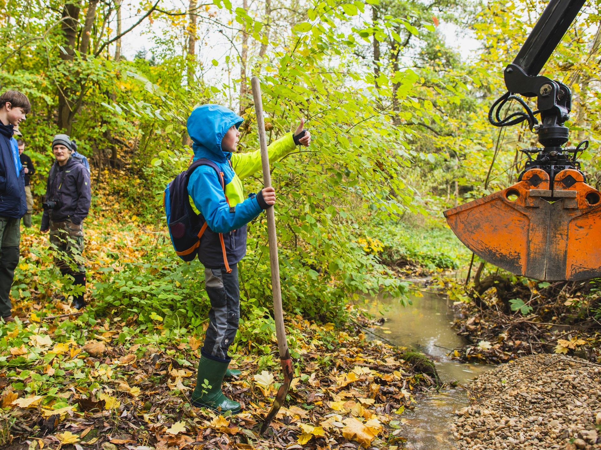 Eine Gruppe von Personen steht an einem kleinen Bach im Wald. Im Vordergrund hält ein Kind einen Holzstab und zeigt mit dem Daumen nach oben, während ein Baggerarm Kies in das Gewässer einbringt. Herbstliches Laub bedeckt den Boden.