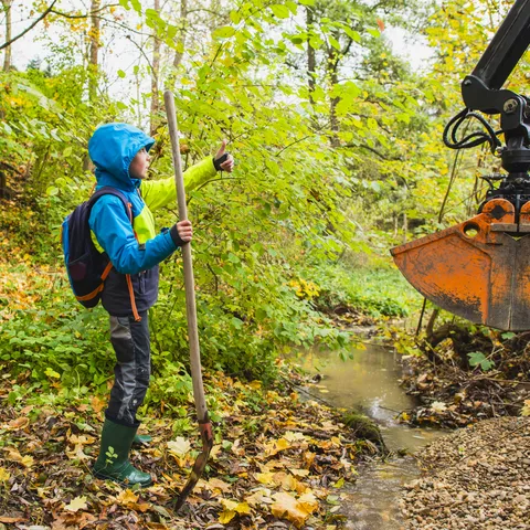 Eine Gruppe von Personen steht an einem kleinen Bach im Wald. Im Vordergrund hält ein Kind einen Holzstab und zeigt mit dem Daumen nach oben, während ein Baggerarm Kies in das Gewässer einbringt. Herbstliches Laub bedeckt den Boden.
