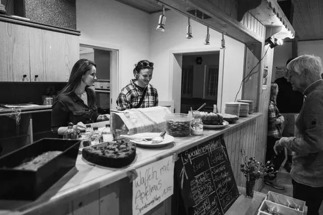 Zwei Personen stehen hinter einer Theke mit Kuchen und Essen, während eine weitere Person davor steht und bedient wird.