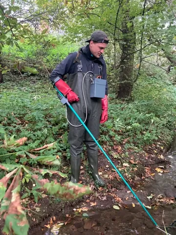 Eine Person steht in Gummistiefeln und Wathose an einem Bach und untersucht das Wasser mit einem langen Messgerät.