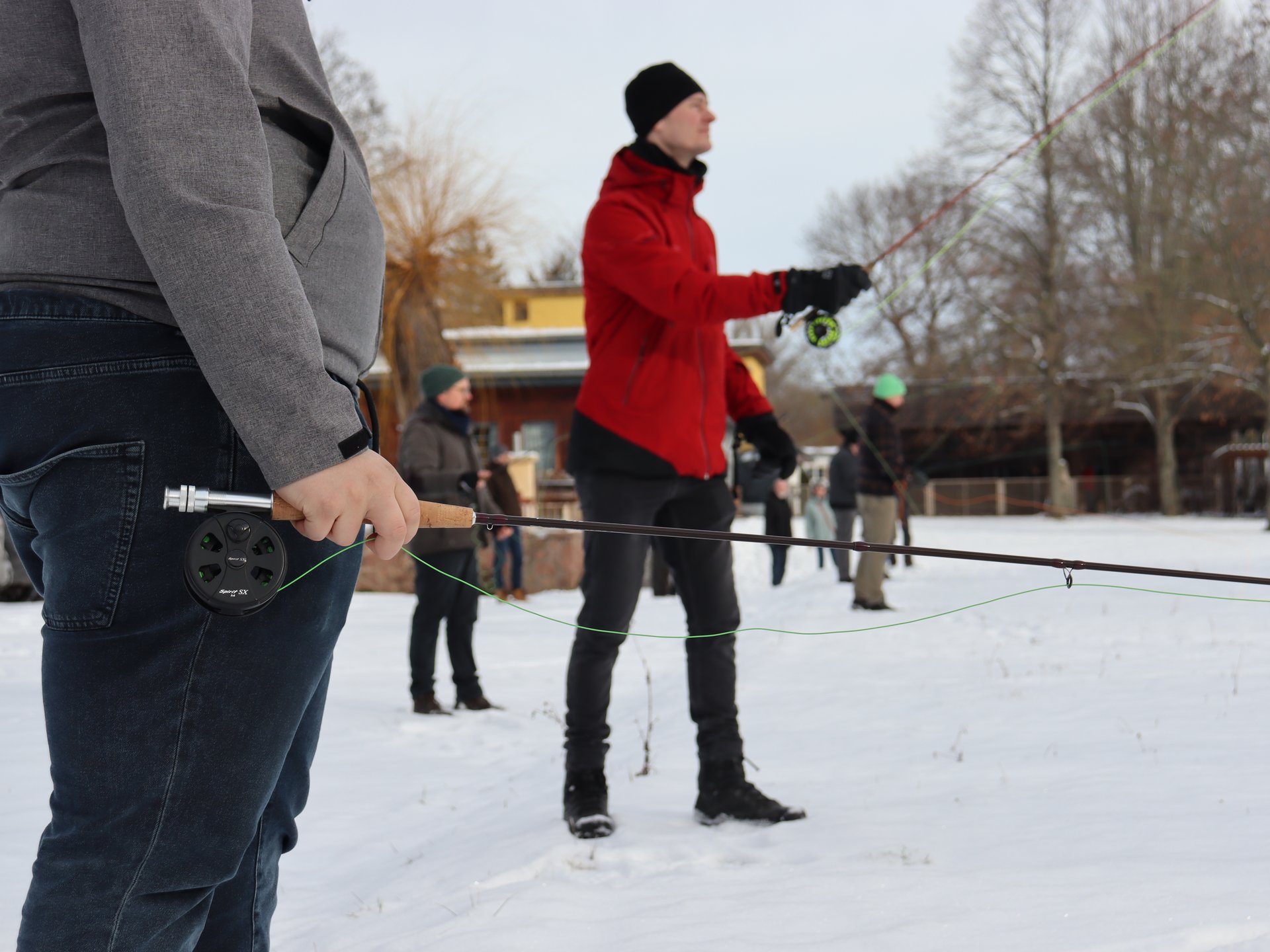 Mehrere Personen üben das Fliegenfischen draußen im Schnee, einige halten Angelruten in den Händen.