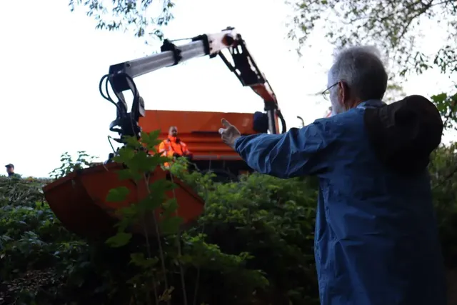 Ein Mann steht vor einem Bagger und hebt seine Arme, während eine Person in orangefarbener Arbeitskleidung in der Baggerschaufel sitzt.