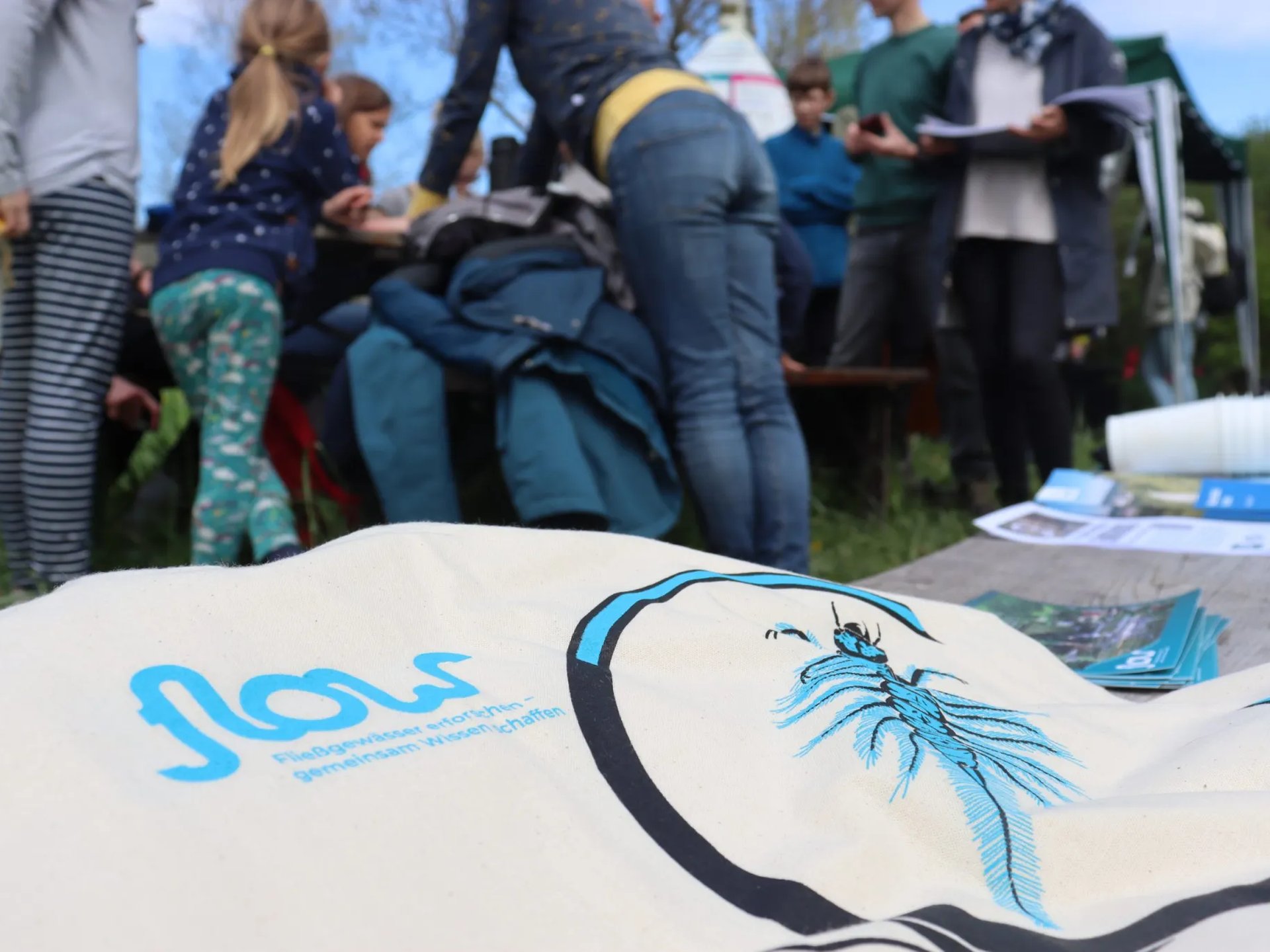 Mehrere Menschen sind im Freien um einen Tisch versammelt, im Vordergrund liegt eine Stofftasche mit einem blauen Wasserinsekt und dem Schriftzug "Flow".