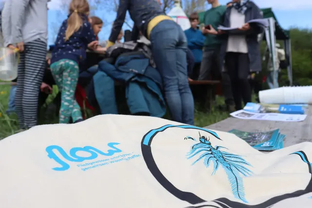 Mehrere Menschen sind im Freien um einen Tisch versammelt, im Vordergrund liegt eine Stofftasche mit einem blauen Wasserinsekt und dem Schriftzug "Flow".