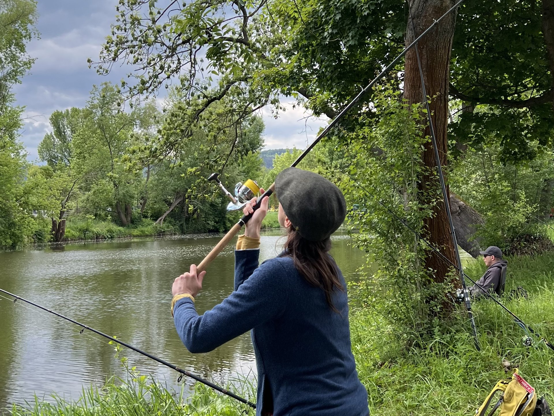 Eine Person steht am Ufer eines Flusses und wirft eine Angelrute aus, umgeben von Bäumen und Gras.