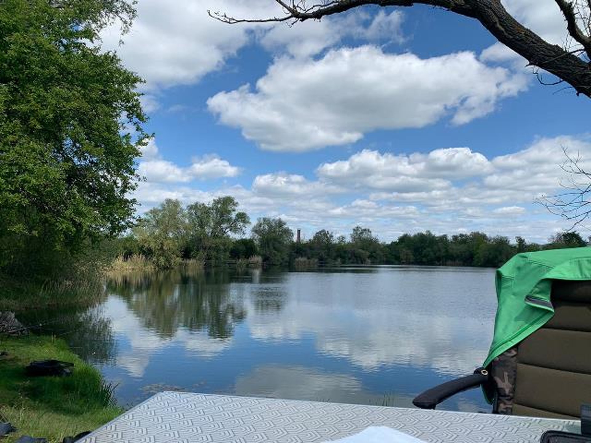 Blick auf einen ruhigen See mit bewölktem Himmel, umgeben von Bäumen, im Vordergrund sind ein Stuhl und ein Tisch am Ufer zu sehen.