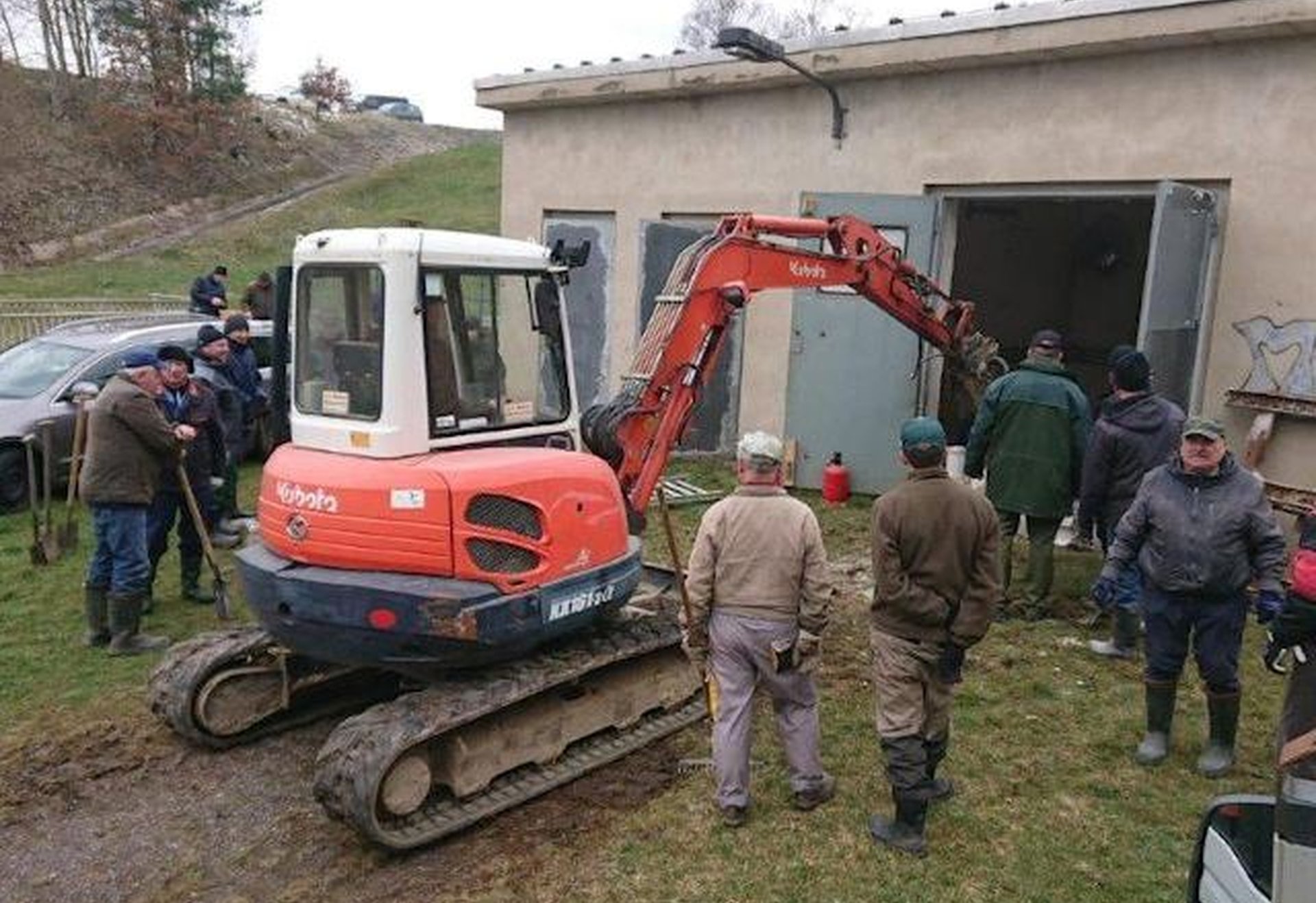 Ein Bagger ist so geparkt, dass sein Greifarm durch eine offene Tür in ein Gebäude hineinragt, während mehrere Personen daneben stehen und zuschauen.